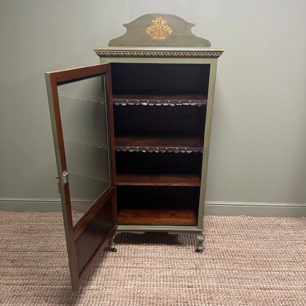 A muted green Edwardian display cabinet featuring a shaped pediment with gilt ornamentation, a single glazed door, and cabriole legs. View X