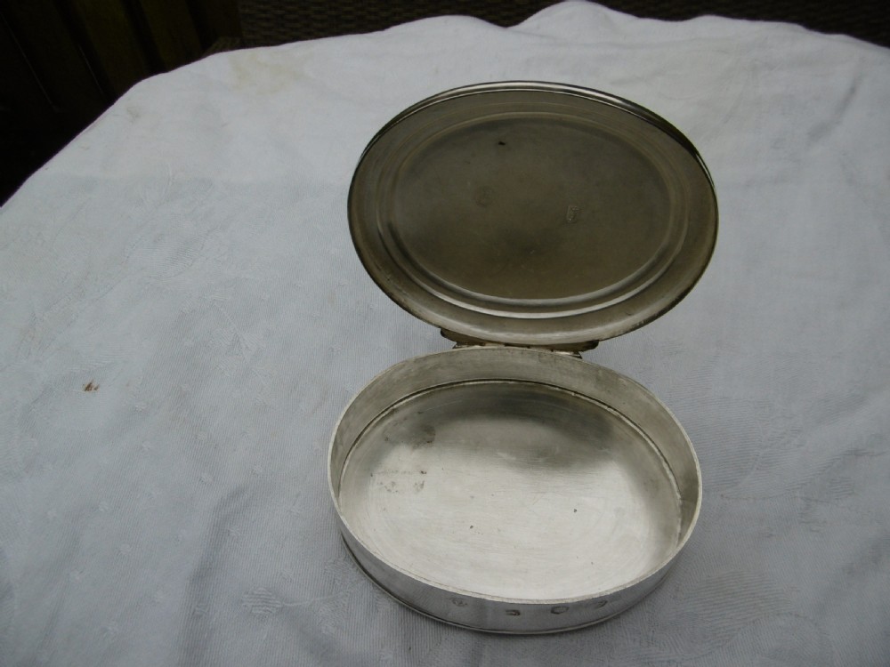 An oval 18th-century sterling silver tobacco box featuring a hinged lid engraved with a central oval crest depicting an open hand beneath a crescent moon. View II