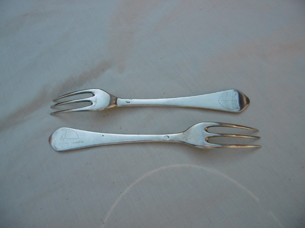 A pair of antique silver table forks featuring three tines and dognose handles engraved with armorial crests.