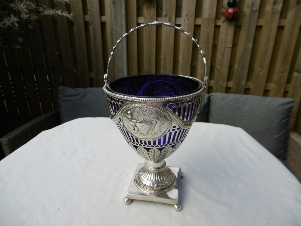 An 18th-century Adam style silver swing-handled basket with a cobalt blue glass liner, resting on a pedestal and square base with ball feet.