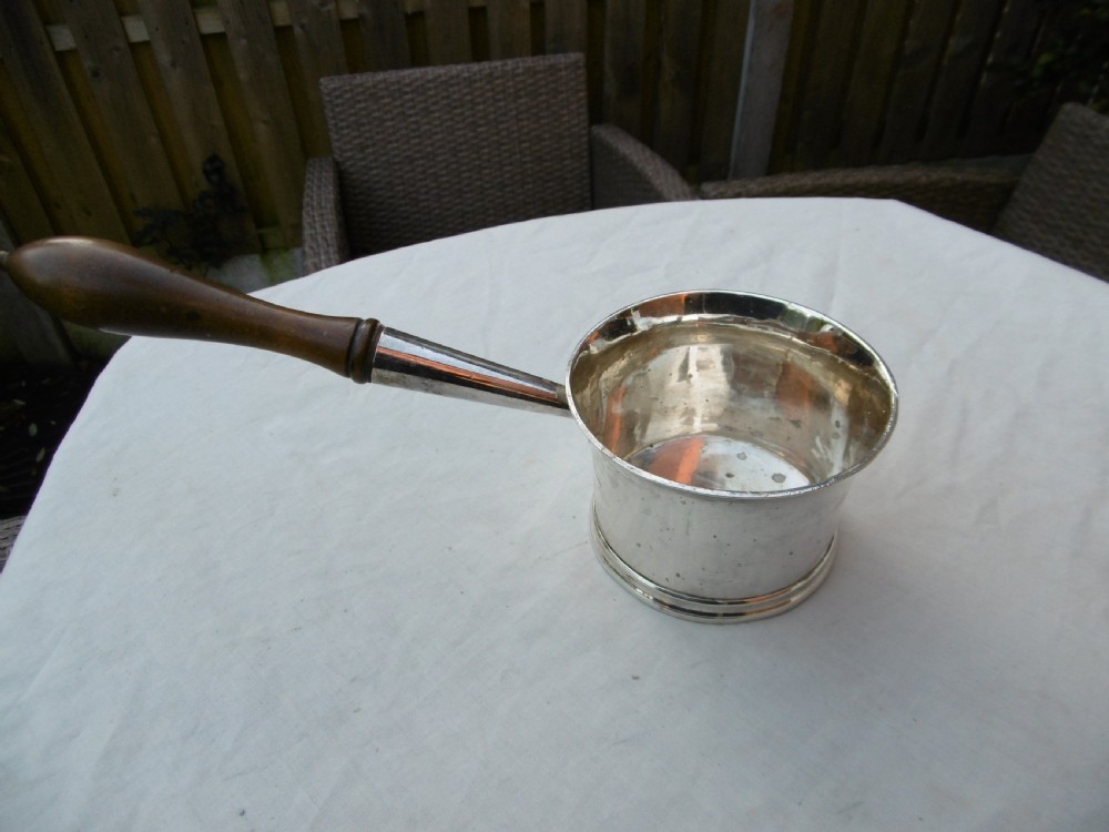 A George I silver brandy pan featuring a flared rim, plain cylindrical body, and a long turned dark wood handle.