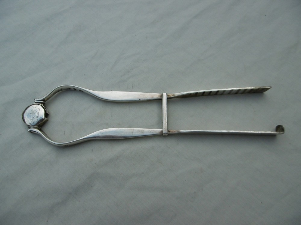A pair of 1799 George III sterling silver serving tongs featuring a circular central boss with an engraved crest, curved arms, and a sliding restraint bar.