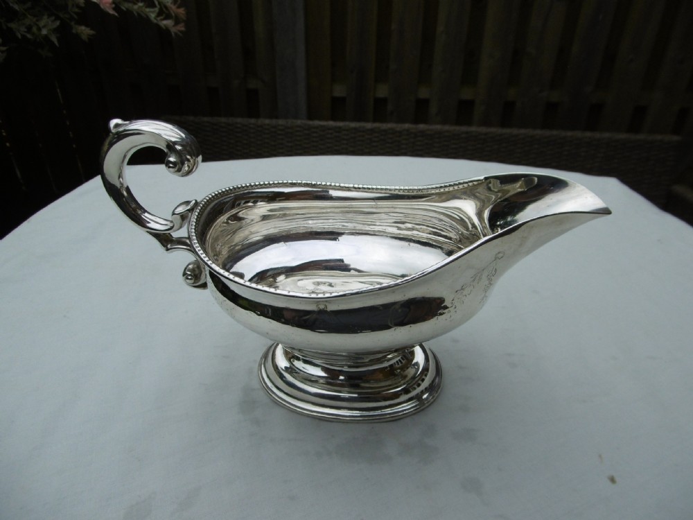 A 1778 Georgian sterling silver sauce boat featuring a beaded rim, a scroll handle, and a oval pedestal foot.