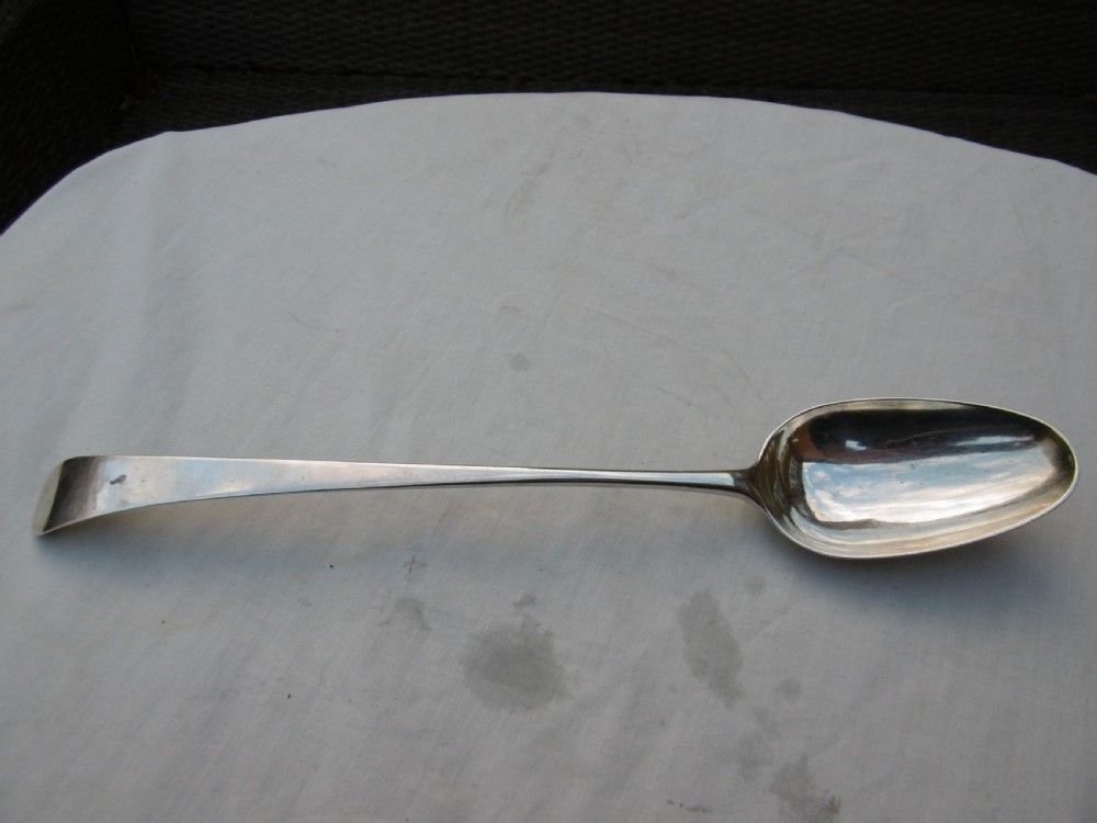 An antique silver basting spoon rests horizontally on a light surface, featuring a long tapered handle and an oval bowl.