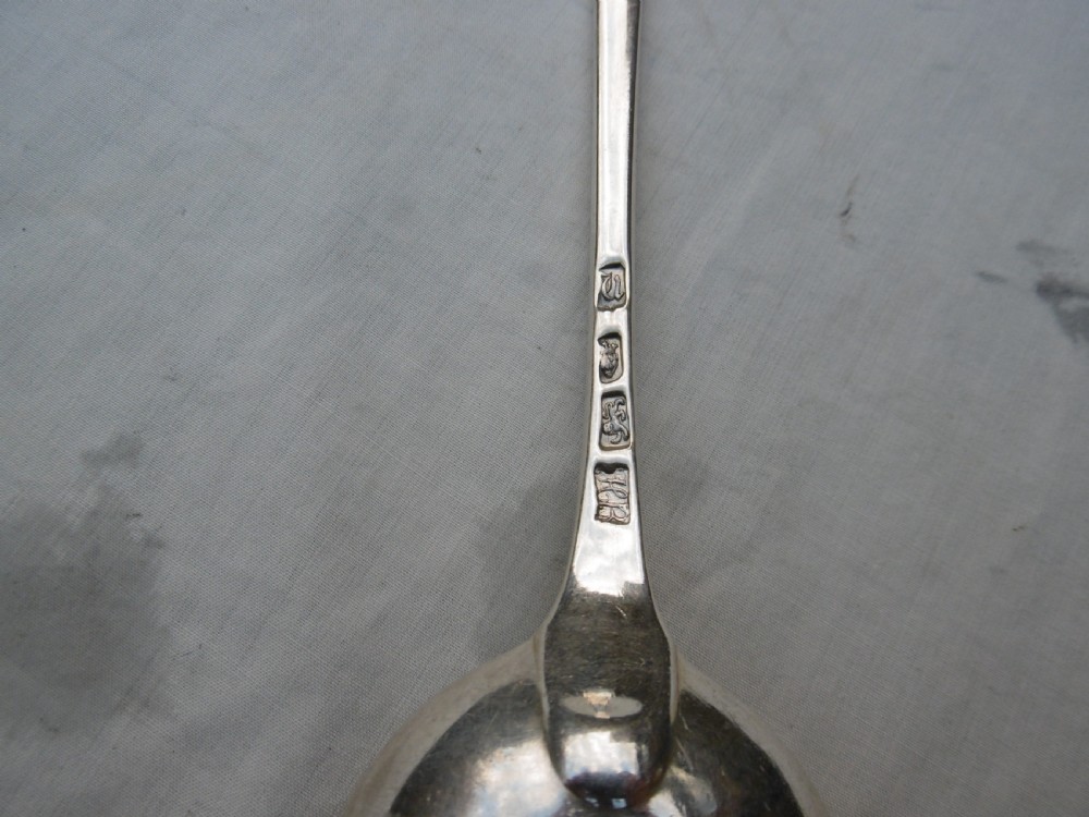 An antique silver basting spoon rests horizontally on a light surface, featuring a long tapered handle and an oval bowl. View II