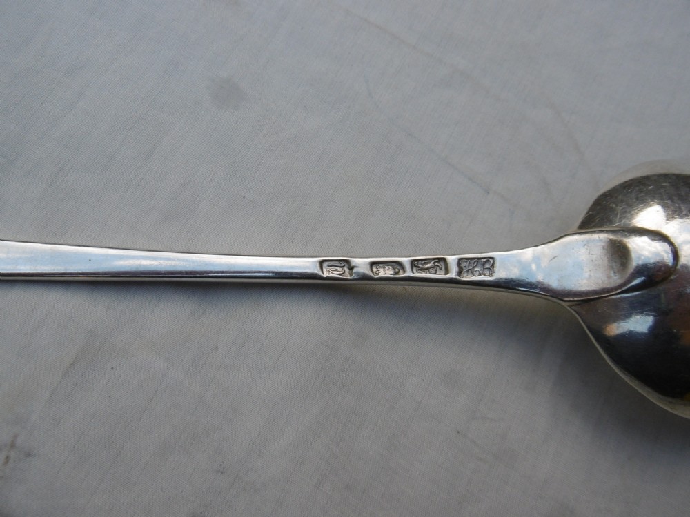 An antique silver basting spoon rests horizontally on a light surface, featuring a long tapered handle and an oval bowl. View III