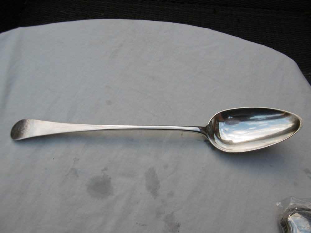 An antique silver basting spoon rests horizontally on a light surface, featuring a long tapered handle and an oval bowl. View IV