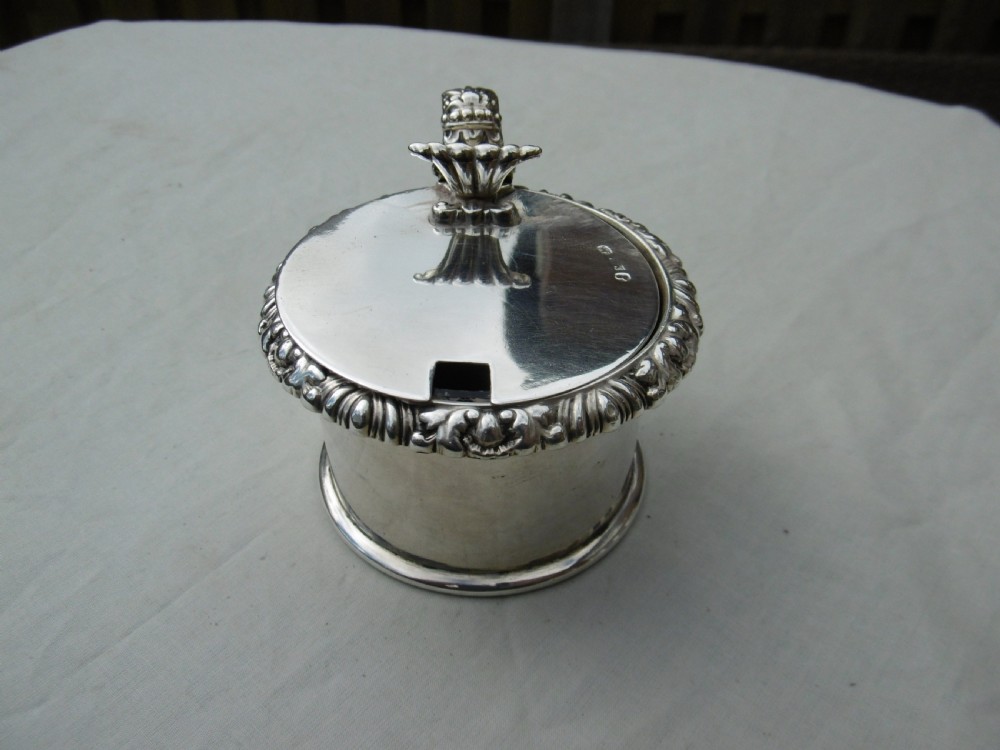 A circular Georgian sterling silver mustard pot featuring a hinged lid with a leaf motif finial and gadrooned border.
