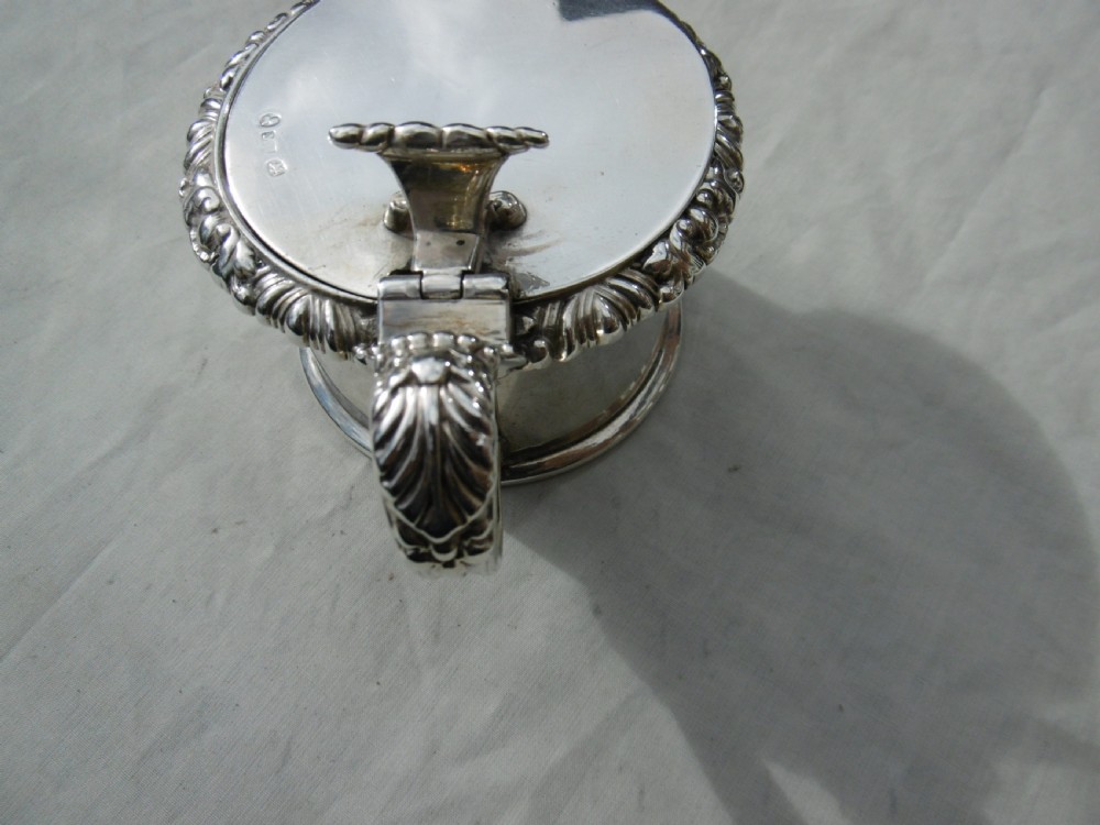 A circular Georgian sterling silver mustard pot featuring a hinged lid with a leaf motif finial and gadrooned border. View IV