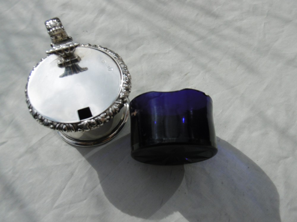 A circular Georgian sterling silver mustard pot featuring a hinged lid with a leaf motif finial and gadrooned border. View V