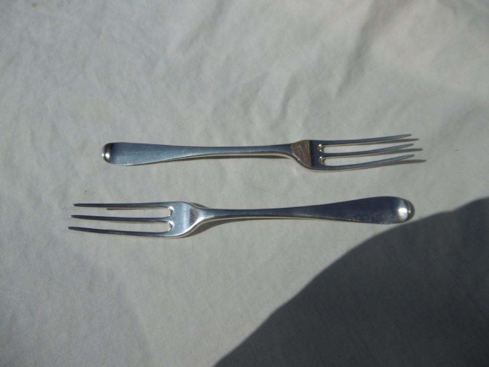 A pair of early 18th-century George I Britannia standard silver three-pronged forks displayed on a white fabric surface.