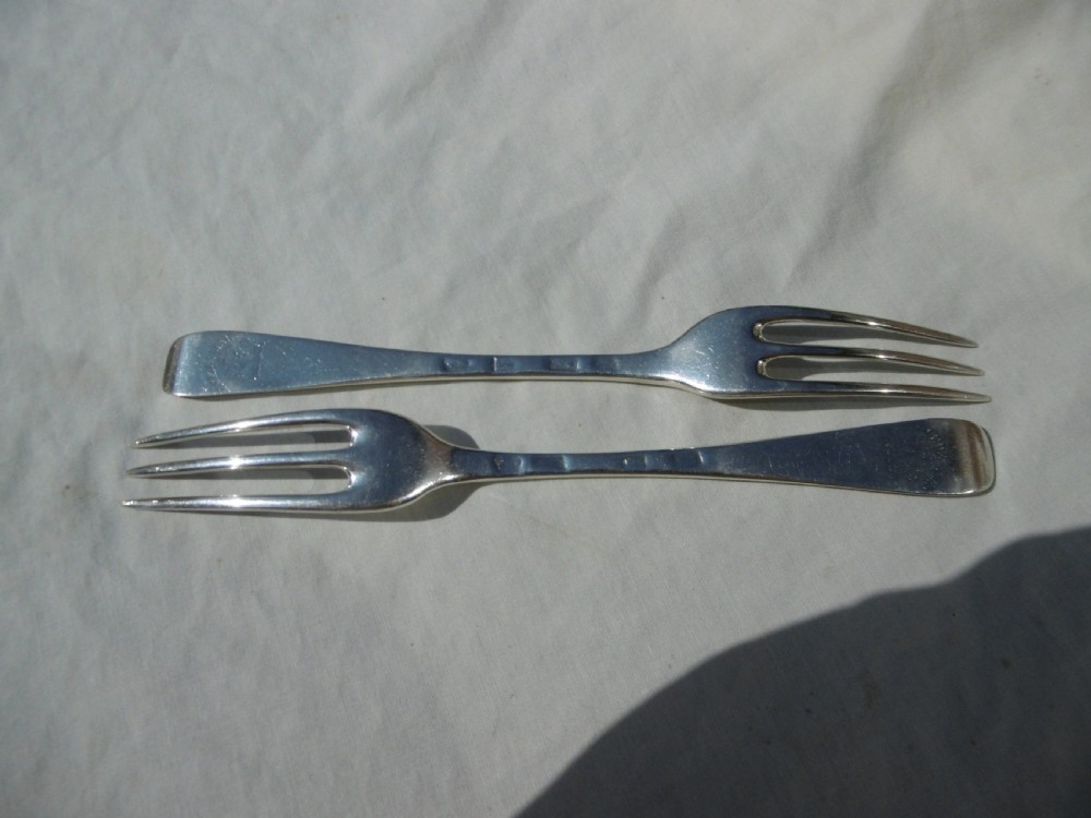A pair of early 18th-century George I Britannia standard silver three-pronged forks displayed on a white fabric surface. View II
