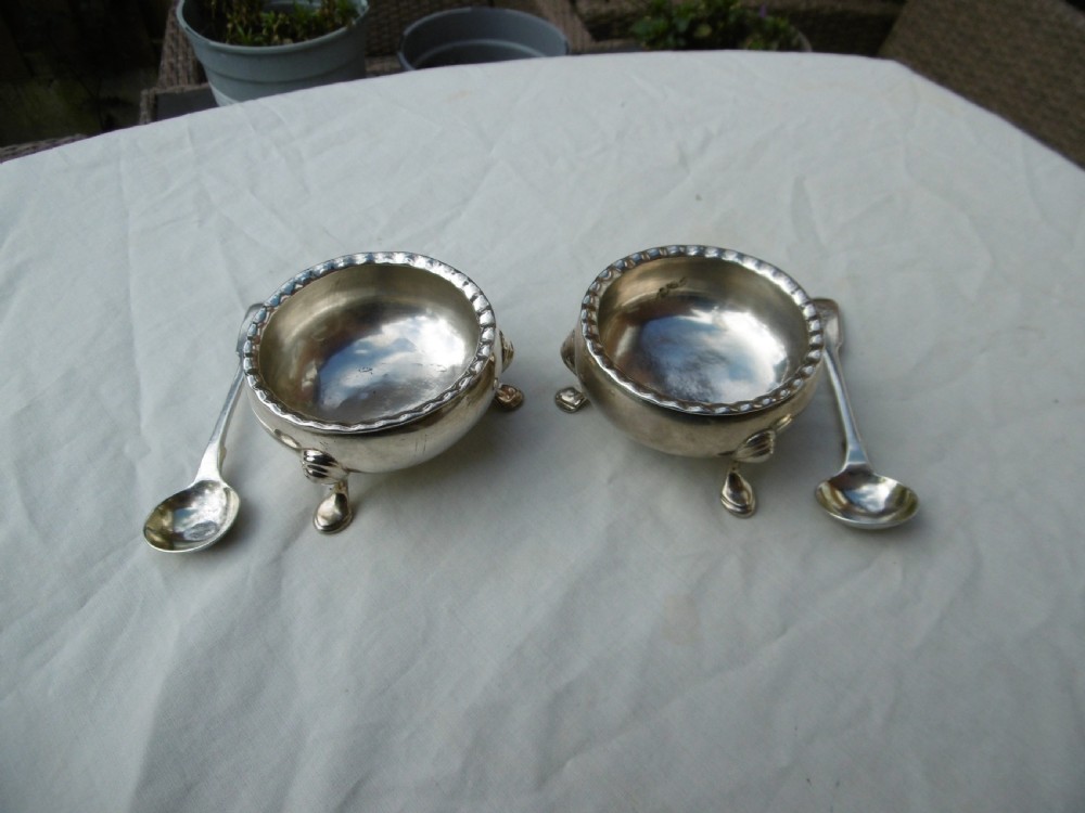 A pair of circular Scottish Georgian sterling silver open salt cellars with hoof feet alongside matching small silver salt spoons.