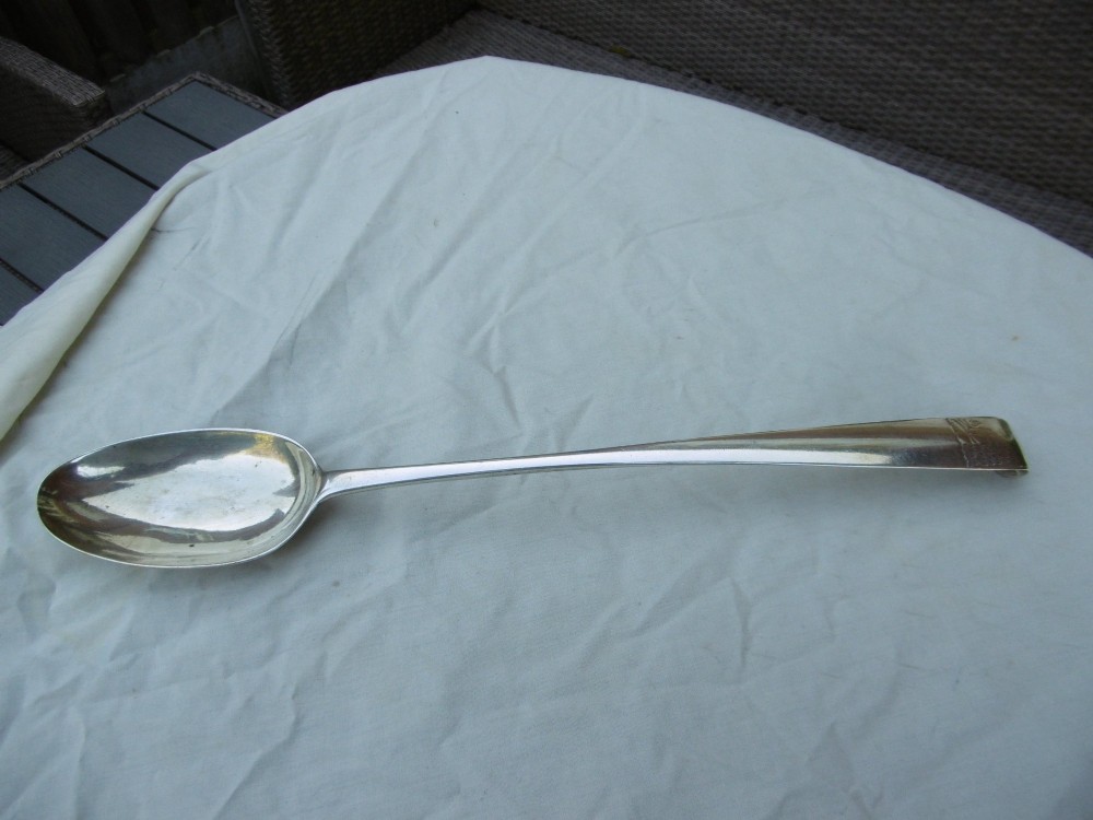 An 18th-century Irish Georgian sterling silver basting spoon featuring a tapered stem with an engraved family crest and a distinctive hooked handle terminal.