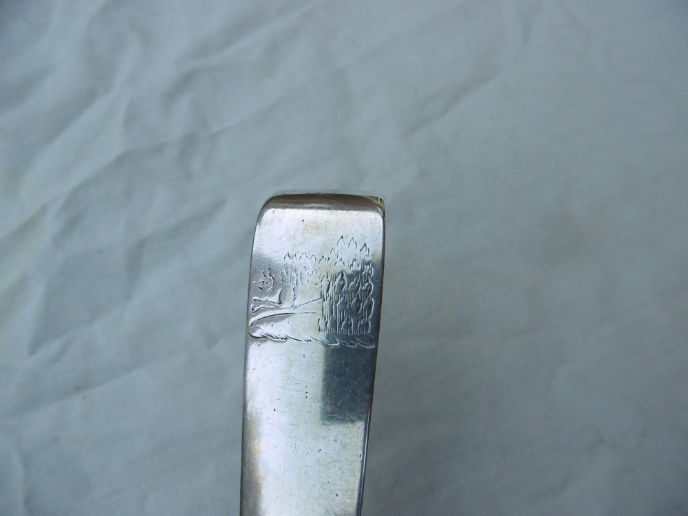An 18th-century Irish Georgian sterling silver basting spoon featuring a tapered stem with an engraved family crest and a distinctive hooked handle terminal. View V