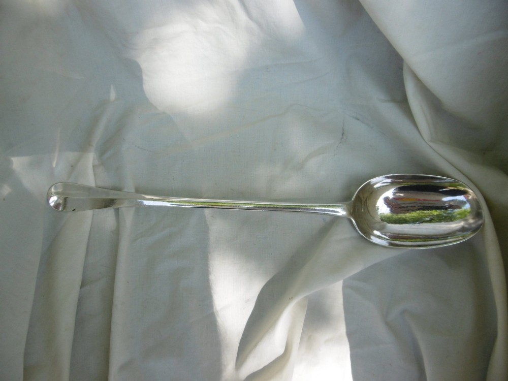 A long George I sterling silver hash spoon featuring an elongated oval bowl resting on white fabric.
