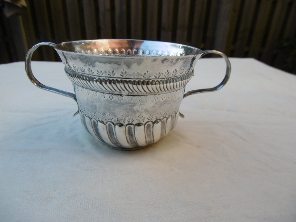 A George I Britannia standard silver porringer with twin scroll handles, fluted lower body decoration, and gadrooned banding.