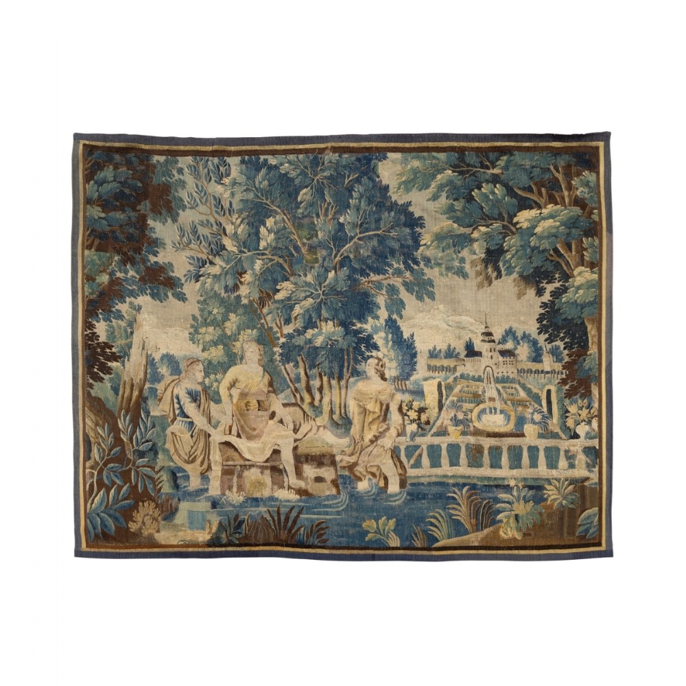 A hand-woven 17th-century Flemish verdure tapestry depicting figures in a classical garden surrounded by dense green trees and distant architectural buildings.