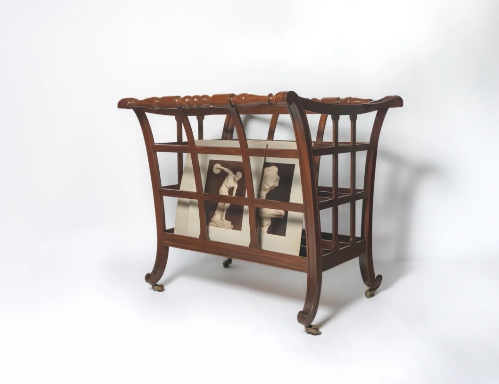 An English Regency mahogany folio stand with open slatted sides, turned top rails, curved legs, and brass castors.