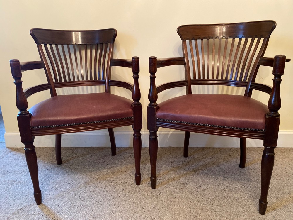 A pair of mahogany club armchairs featuring vertical slat backrests, curved arms with turned supports, and reddish-brown leather upholstered seats with stud detailing.