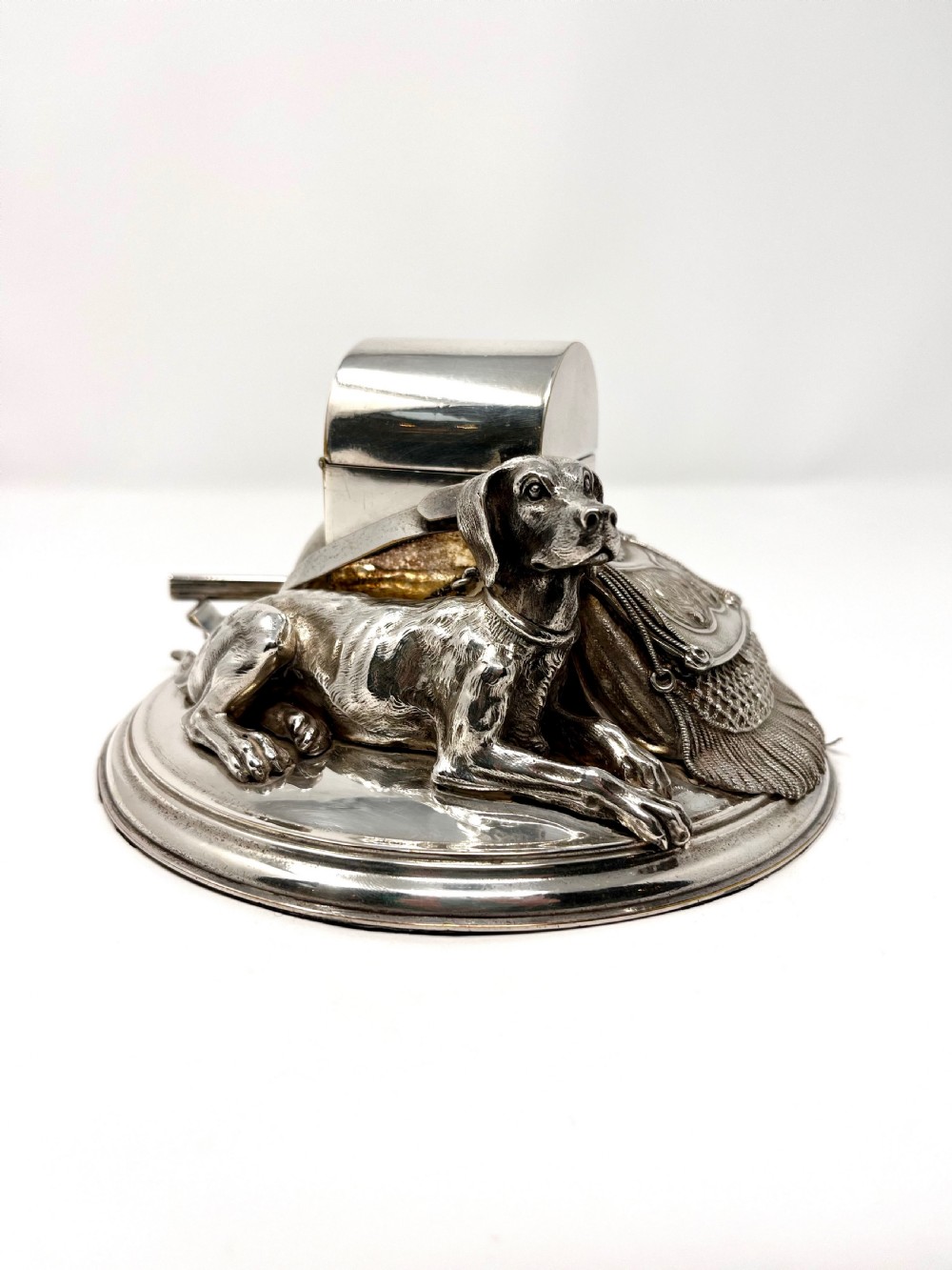 A Victorian silver-plated desk inkstand featuring a sculpted reclining hunting hound next to a game bag and domed ink container on a circular stepped base. View VI
