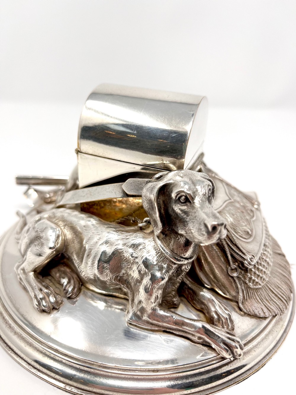 A Victorian silver-plated desk inkstand featuring a sculpted reclining hunting hound next to a game bag and domed ink container on a circular stepped base. View II