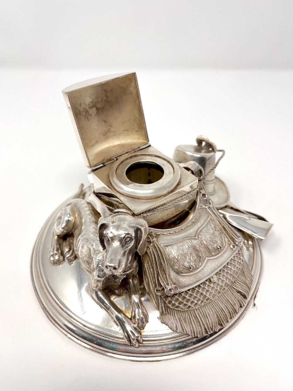 A Victorian silver-plated desk inkstand featuring a sculpted reclining hunting hound next to a game bag and domed ink container on a circular stepped base. View V