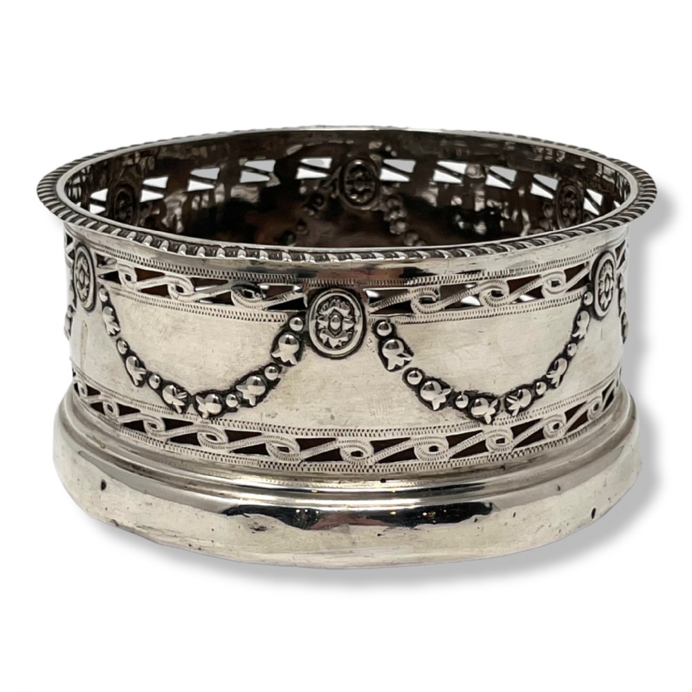 A circular silver bottle coaster featuring intricate pierced fretwork, floral swags, and oval medallions around the body.