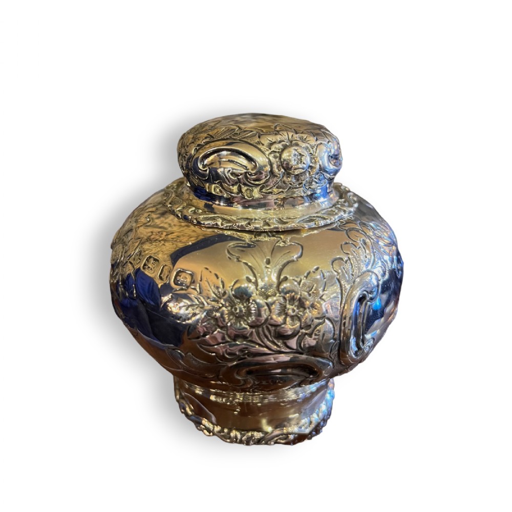A bulbous antique silver tea caddy featuring hand-chased floral repousse decoration, visible hallmarks, and a fitted domed cap.