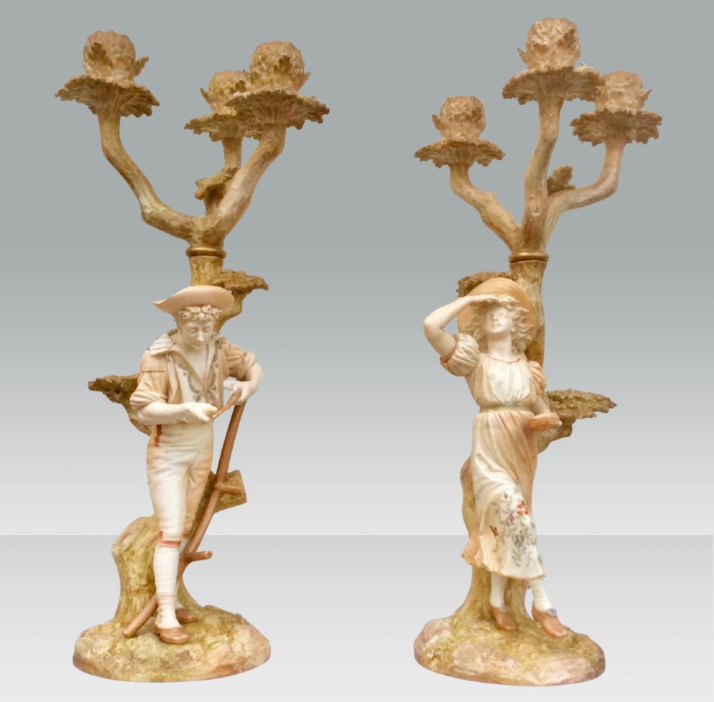 A pair of antique Royal Worcester porcelain candelabra featuring finely modelled male and female rustic figures standing before tree trunk candle branches.