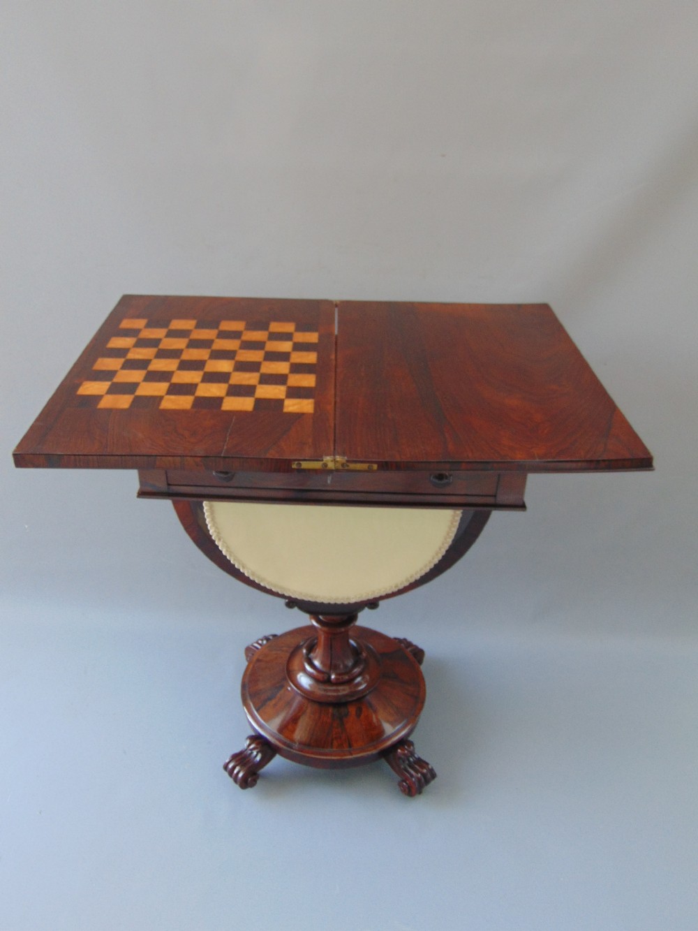 A William IV rosewood sewing and games table with a fold-over top, single drawer, suspended cream fabric work basket, and turned pedestal base with scroll feet. View II