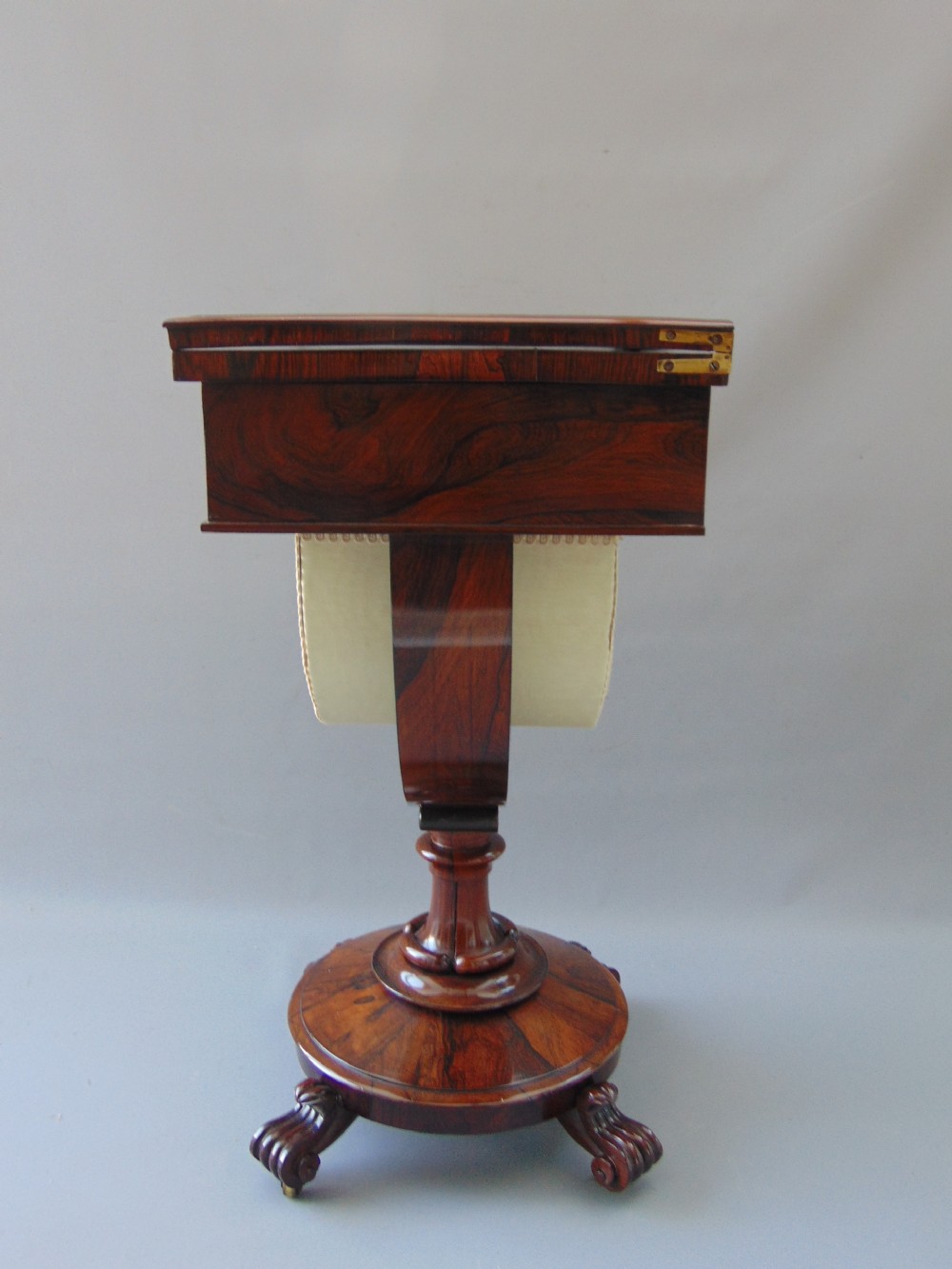 A William IV rosewood sewing and games table with a fold-over top, single drawer, suspended cream fabric work basket, and turned pedestal base with scroll feet. View IV