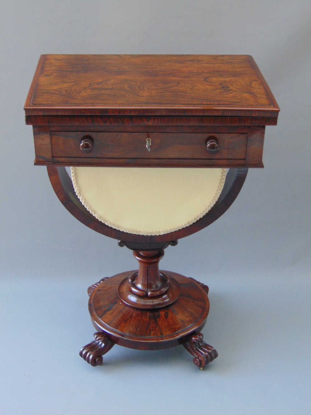A William IV rosewood sewing and games table with a fold-over top, single drawer, suspended cream fabric work basket, and turned pedestal base with scroll feet. View IX