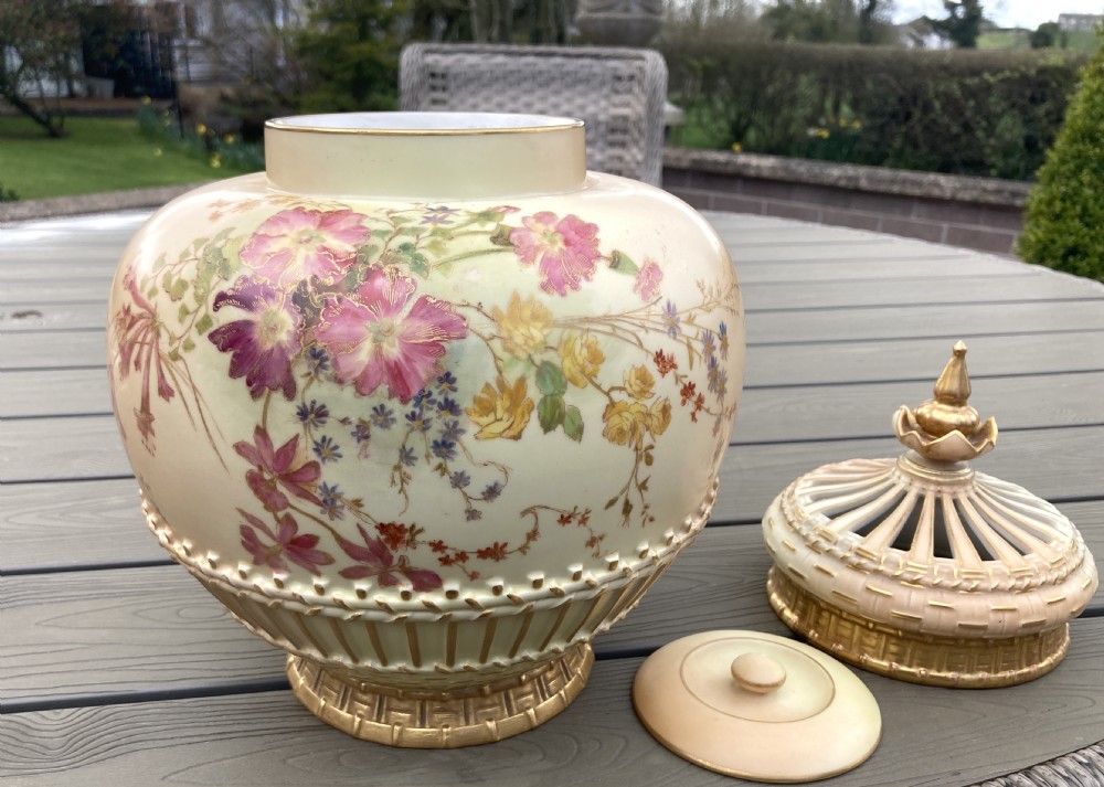 A bulbous Royal Worcester blush ivory porcelain pot pourri vase featuring hand-painted floral blooms, basketweave moulded relief details, and a decorative finial cover. View V