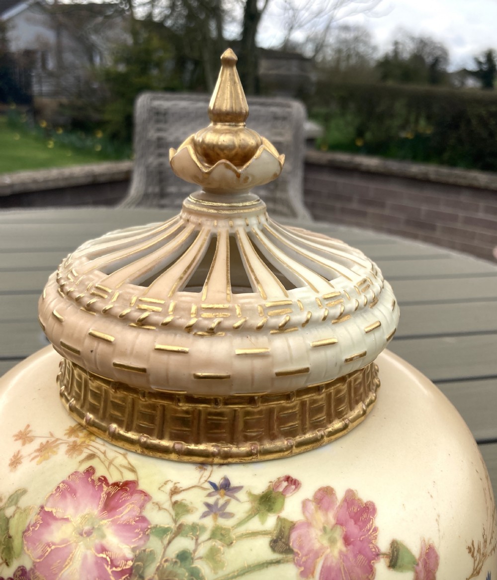 A bulbous Royal Worcester blush ivory porcelain pot pourri vase featuring hand-painted floral blooms, basketweave moulded relief details, and a decorative finial cover. View VII