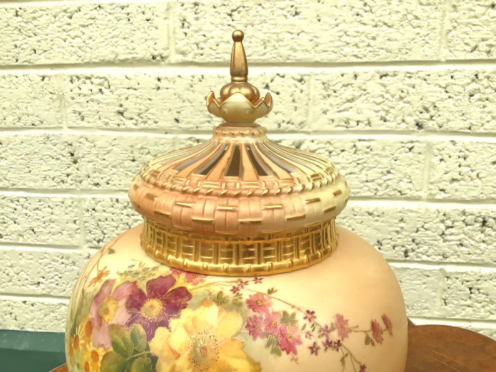 A globular Royal Worcester blush ivory pot pourri vase featuring hand-painted floral motifs, basketweave relief detail, and a pierced gilded lid. View V