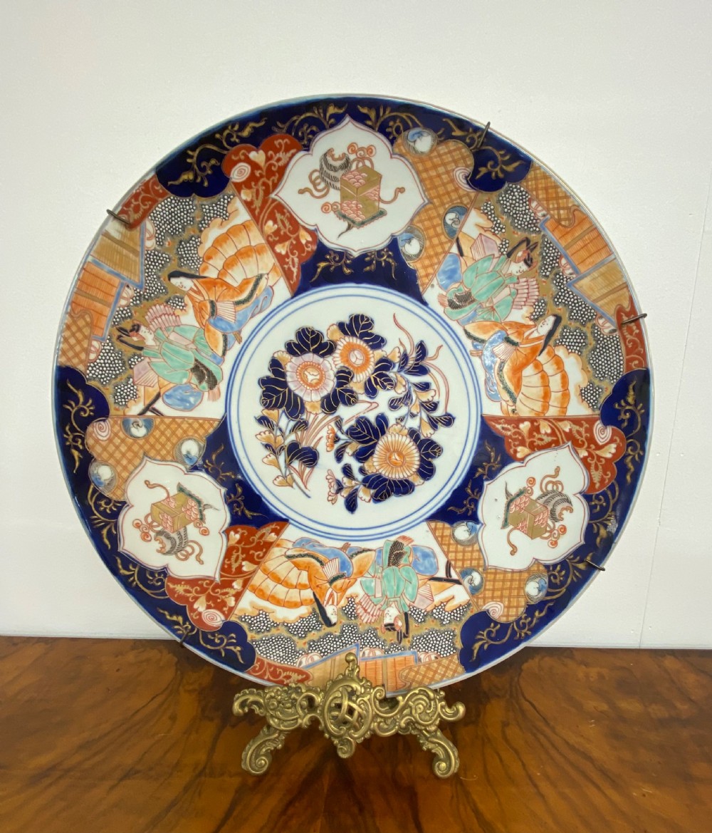 A circular antique Japanese Imari porcelain charger plate decorated with blue, orange, green, and gold panelled designs featuring figures and botanical motifs.