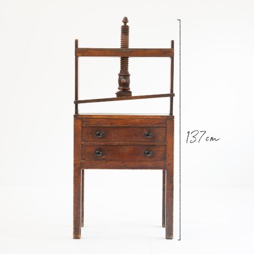 fig 1. A Victorian mahogany and pine linen press featuring a central wooden screw mechanism over two small drawers with turned black knobs and four square legs.
