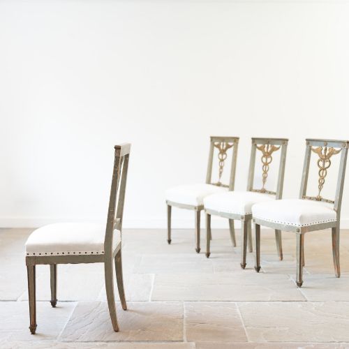 fig 1. A set of four 19th-century Northern European Neoclassical side chairs featuring distressed grey paintwork, off-white linen upholstery, and gilded caduceus motifs in the open back frames.
