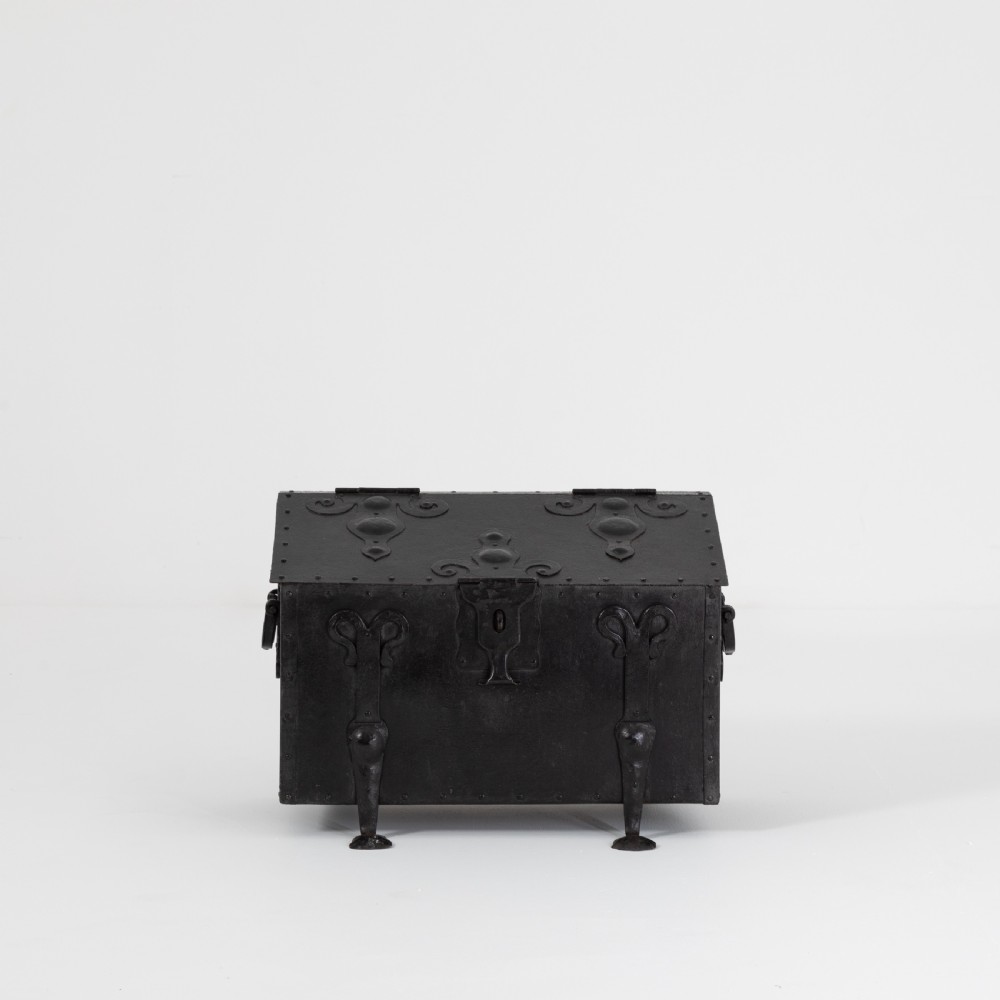A heavy black wrought iron chest featuring a gabled hinged lid, decorative strap hinges, rivets, and stylised scrollwork appliques.