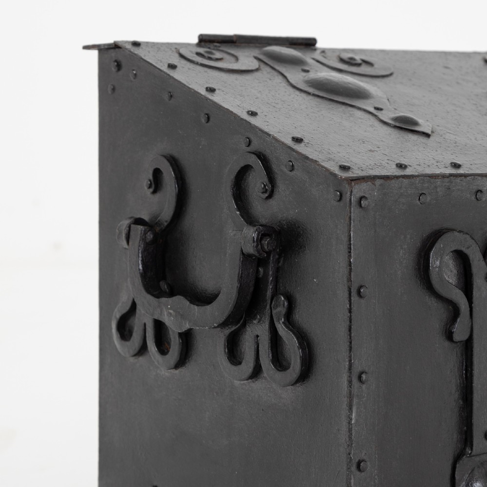 A heavy black wrought iron chest featuring a gabled hinged lid, decorative strap hinges, rivets, and stylised scrollwork appliques. View IV
