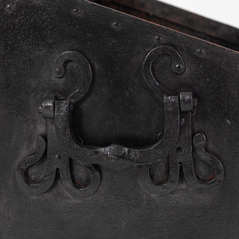 A heavy black wrought iron chest featuring a gabled hinged lid, decorative strap hinges, rivets, and stylised scrollwork appliques. View V