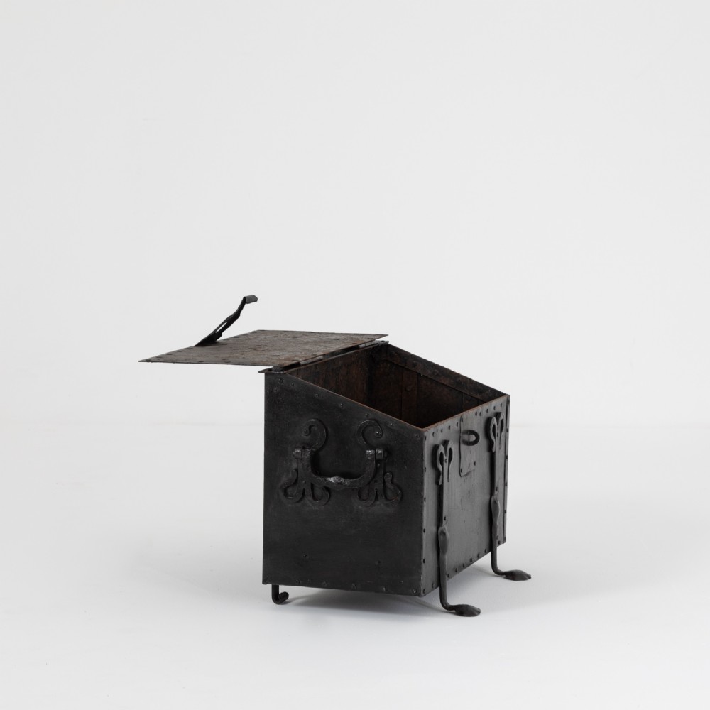 A heavy black wrought iron chest featuring a gabled hinged lid, decorative strap hinges, rivets, and stylised scrollwork appliques. View VI