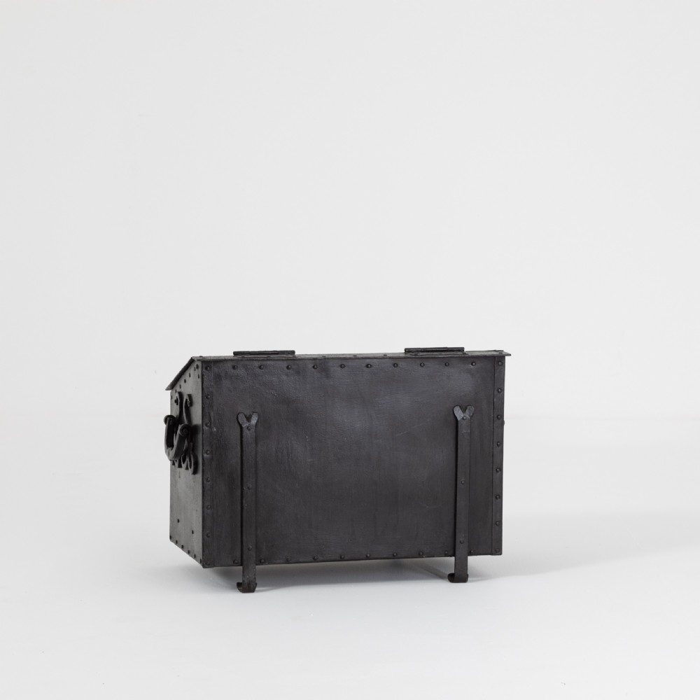 A heavy black wrought iron chest featuring a gabled hinged lid, decorative strap hinges, rivets, and stylised scrollwork appliques. View VII