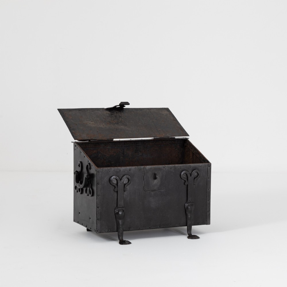 A heavy black wrought iron chest featuring a gabled hinged lid, decorative strap hinges, rivets, and stylised scrollwork appliques. View VIII