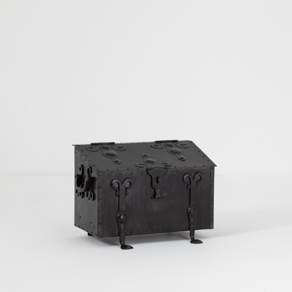 A heavy black wrought iron chest featuring a gabled hinged lid, decorative strap hinges, rivets, and stylised scrollwork appliques. View IX