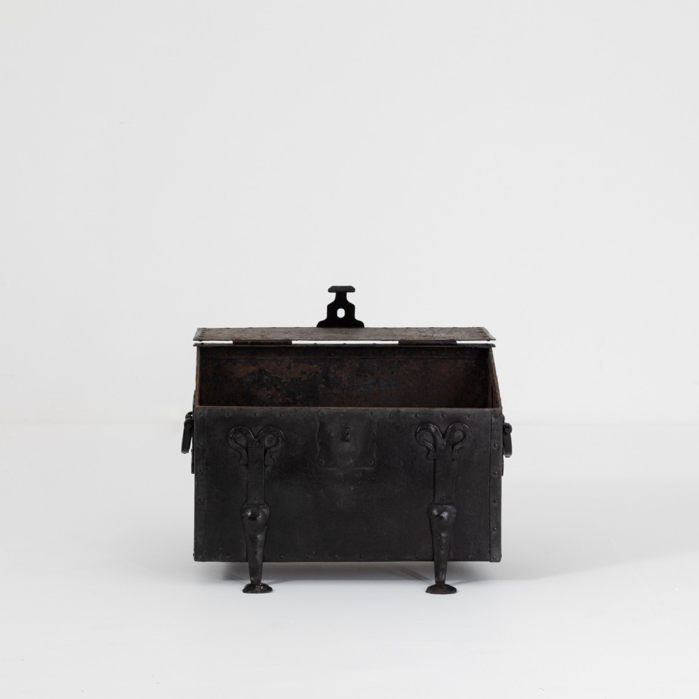 A heavy black wrought iron chest featuring a gabled hinged lid, decorative strap hinges, rivets, and stylised scrollwork appliques. View X