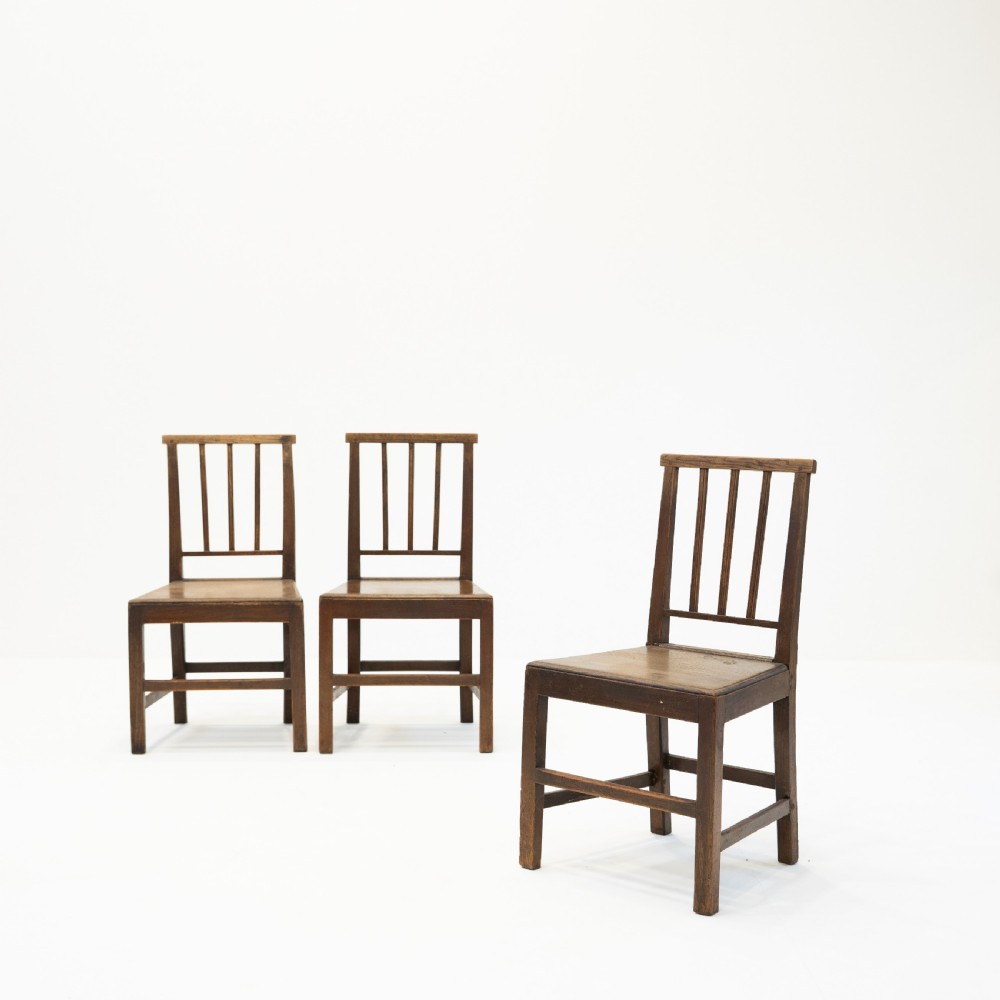A set of three 18th-century dark wooden farmhouse chairs featuring vertical slat backs, solid wooden seats, and straight legs connected by stretchers.