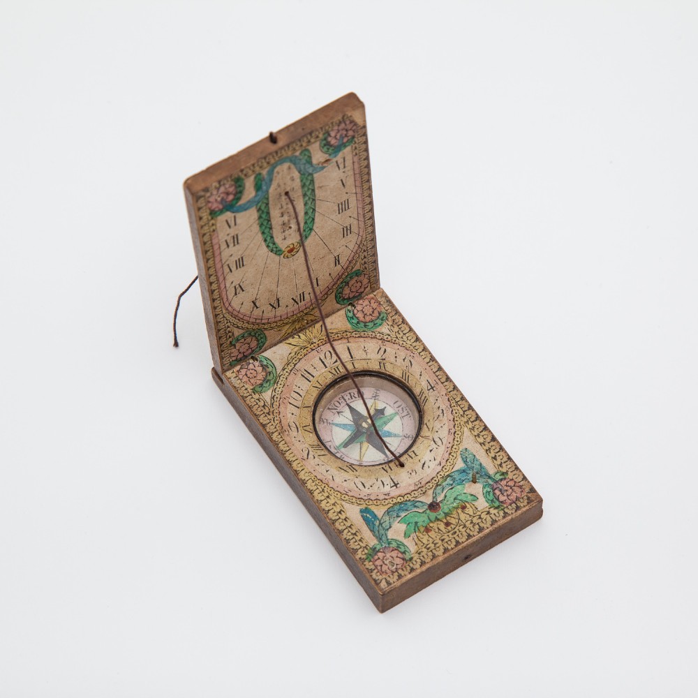 A small hinged rectangular wooden diptych sundial displayed open, showing hand-tinted paper hour dials, floral details, a central thread gnomon, and a glass-covered circular compass.