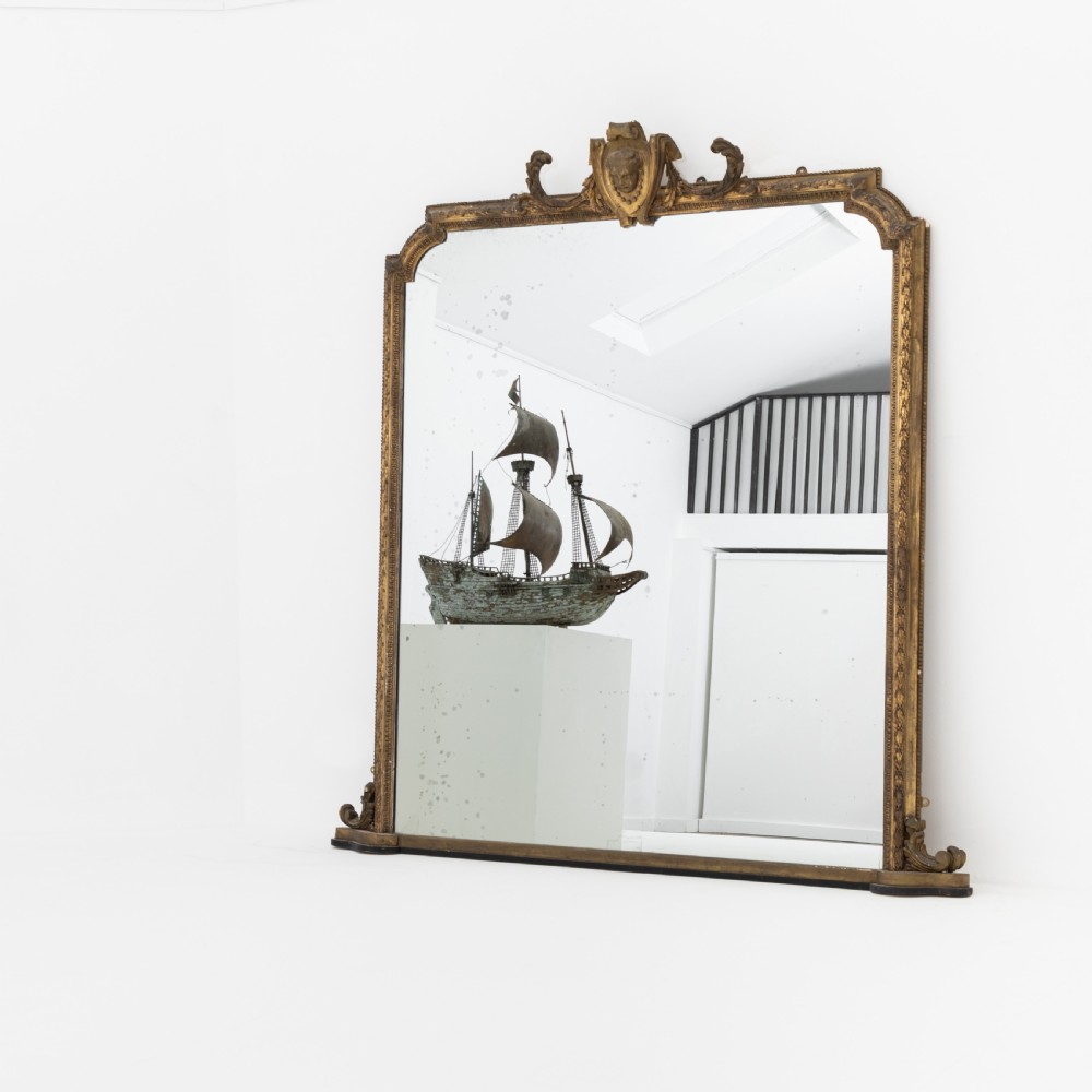 A large Victorian giltwood overmantel mirror featuring an arched frame, detailed cresting with a carved mask, and ornate scroll base feet.